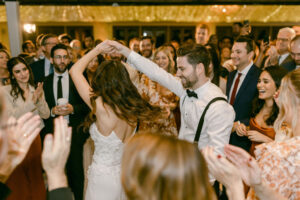 Groom twirls his bride during their first dance while surrounded by their loved ones on the dance floor.