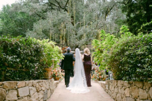 Bride walks down the aisle to her groom, arm in arm with parents on either side.