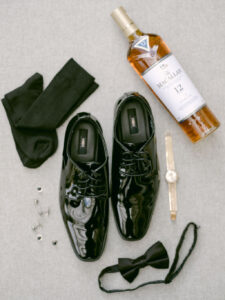 Flatlay of groom's wedding day details including his shoes, socks, cufflinks, bow tie, watch and a bottle of whiskey.