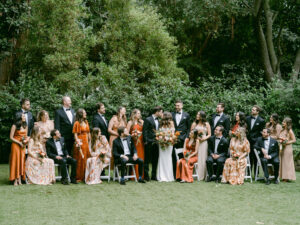 Full bridal party photo as bride and groom kiss and the bridal party smile toward the happy couple. Fall toned bridal party attire.