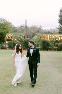 Groom holds bride's train as they smile at each other and share a walk.