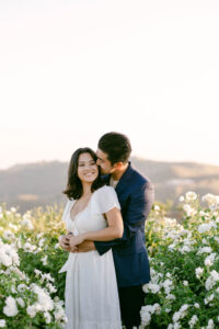 Groom wraps his arms around his new fiance and kisses her cheek as they stand in the middle of the white rose garden at Stonewall Ranch Malibu following their beautiful proposal.