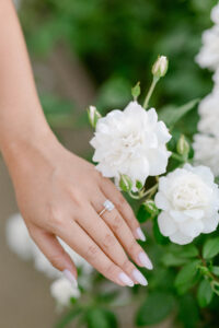 Image shows emerald cut engagement ring on newly engaged woman's finger next to the beautiful white roses at Stonewall Ranch.