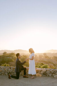 Asian groom wearing a blue suit get's down on one knee as he proposes to his bride to be wearing a flowy white dress at sunset in the hills of Malibu.