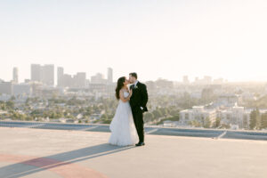 Bride and groom share a kiss on the helipad of the Four Seasons Beverly Hills with DTLA in the background as the sun sets.
