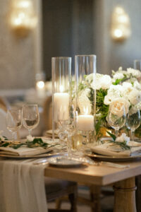 Cream colored, chiffon table runner flows off the end of the light wood tables, decorated with white candles and white floral centerpieces.