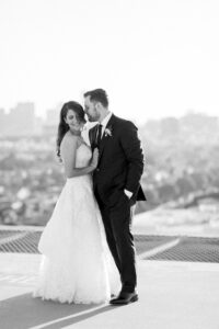 Black and white photo of bride smiling over her shoulder as her groom pulls her close and whispers into her ear on the rooftop of the Four Seasons Beverly Hills.