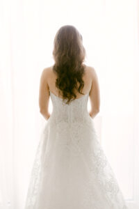 Bride looks out the window toward her wedding ceremony at the Four Seasons Beverly Hills. Showcasing the back of her lace, strapless, Monique Lhuillier wedding gown.