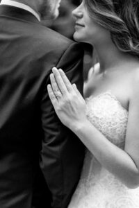 Close up, black and white photo of bride and groom looking at each other, as bride rests her hand on his arm, showing off her wedding ring.