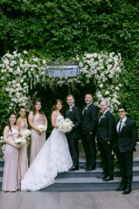 Timeless wedding party portrait in front of white floral chuppah. Bride and groom stand together in the center and their bridesmaids and groomsmen stand next to them on the steps.