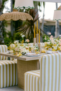 A table surrounded by yellow and white striped chairs, adorned with a centerpiece of white and yellow flowers, yellow candles with wooden candle holders and hanging wicker and fabric lights.